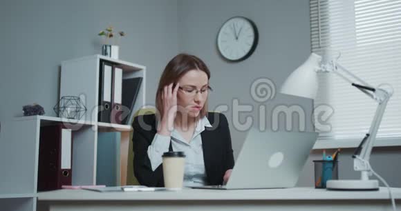 年轻疲惫的女人戴着眼镜坐在办公室里的笔记本电脑前几乎睡着了视频的预览图