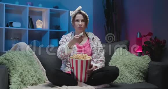 性格开朗的休闲女士带着滑稽的脸吃爆米花和看电影坐在沙发上享受真诚的积极情绪视频的预览图
