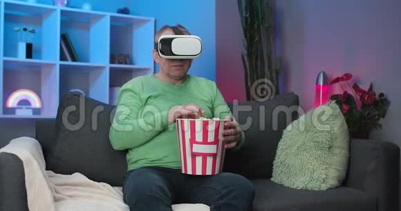老人戴着虚拟现实眼镜在家坐在沙发上吃爆米花看电影视频的预览图