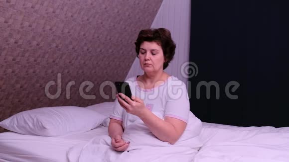 成年妇女坐在床上睡觉后使用电话视频的预览图