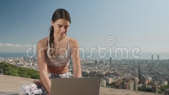 女人坐在莲花姿势与笔记本电脑户外城里的女孩在电脑上打字视频的预览图