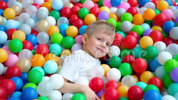 金发小男孩躺在多种颜色的塑料球在大干划水池在游戏中心对着镜头微笑视频的预览图