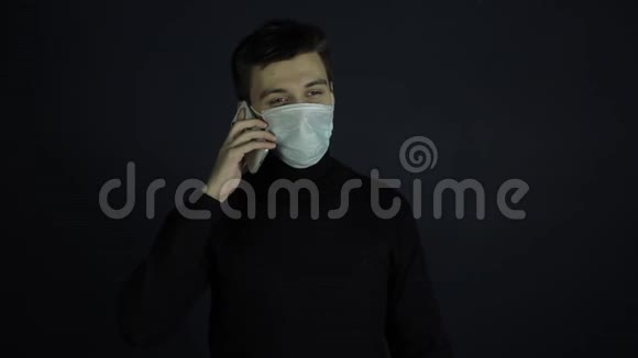 一个年轻的白种人戴着一个防护卫生的医疗面罩在电话里说话冠状病毒保护室内视频的预览图