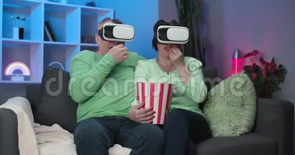 老白种人配偶一起坐在沙发上吃爆米花在VR护目镜上看电影一对夫妇坐着视频的预览图