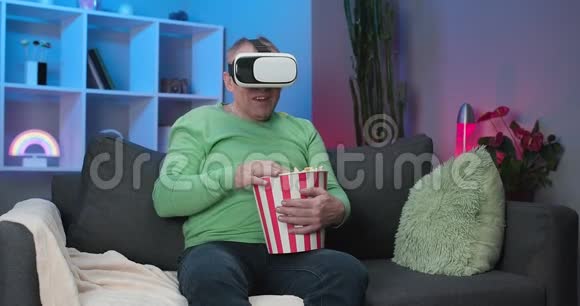 高加索人用虚拟现实耳机看电视坐在沙发上在房间里吃爆米花视频的预览图