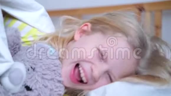 可爱的金发小女孩笑着躺在白色的床下被子女孩拥抱泰迪熊早上好4k个镜头视频的预览图