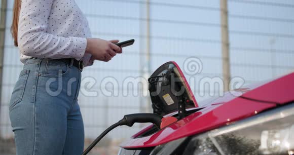 汽车电气充电过程即将结束白种人女孩使用智能手机和等待电源连接到视频的预览图