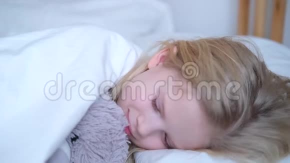 一大早金发小女孩在床上醒来对着镜头微笑女孩抱着一只泰迪熊与上床视频的预览图