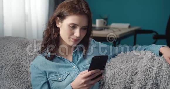 微笑的年轻女士坐在沙发上用智能手机视频的预览图