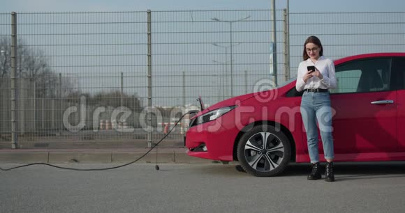 女孩站在她的红色电动汽车附近的电话等待车辆什么时候充电生态汽车连接视频的预览图