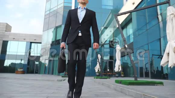 年轻的商人穿着黑色西装公文包沿着现代办公楼走着自信的男性企业家视频的预览图