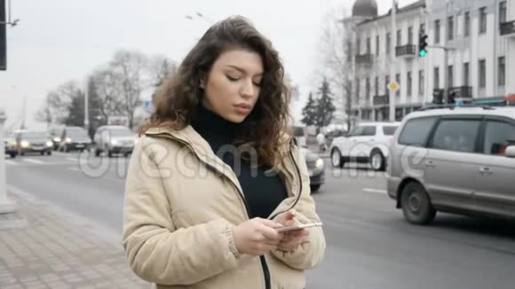 在繁忙的城市道路上一个头发卷曲的美丽黑发女孩在她的智能手机上写着一条信息视频的预览图