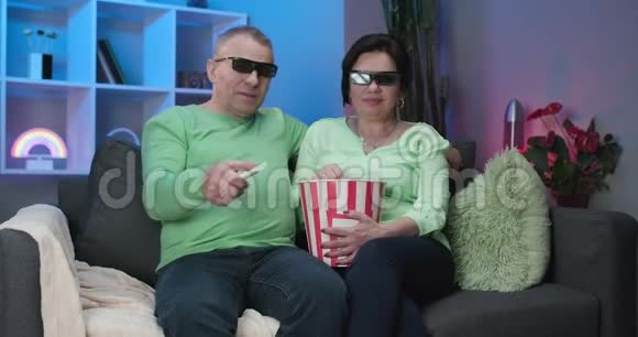 年长夫妇戴着3D眼镜吃爆米花年长的男人和女人一起看电视坐在家里的沙发上视频的预览图