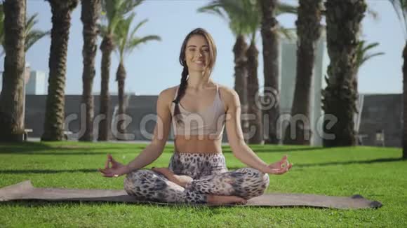 微笑瑜伽教练坐在公园的垫子上在户外摆着摄像机的女孩视频的预览图