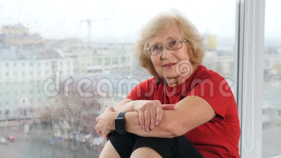 穿着t恤看镜头的退休妇女的肖像坐在窗台上看着老人和蔼的养老老人视频的预览图