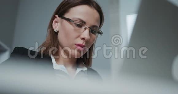 一位戴着眼镜精力充沛的女士坐在办公桌前的时候她正在用笔记本电脑拍摄她的特写镜头视频的预览图