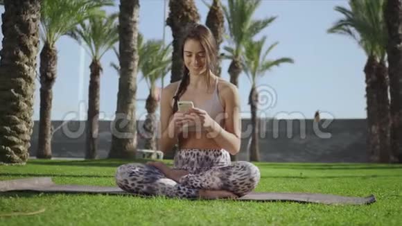 女人坐在莲花姿势与智能手机公园里用手机的女孩视频的预览图
