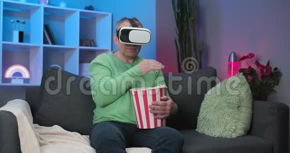 老人坐在沙发上拿着爆米花他戴着虚拟现实眼镜举起手选择性地看着视频的预览图