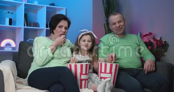高白种人夫妇和5岁的孙女坐在家里的沙发上微笑着看电视吃爆米花视频的预览图