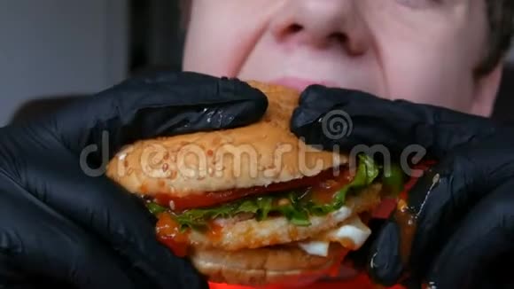 一个十几岁的孩子吃了一个大的多汁汉堡里面有一个鸡蛋一个西红柿沙拉和红酱双手戴黑色手套视频的预览图