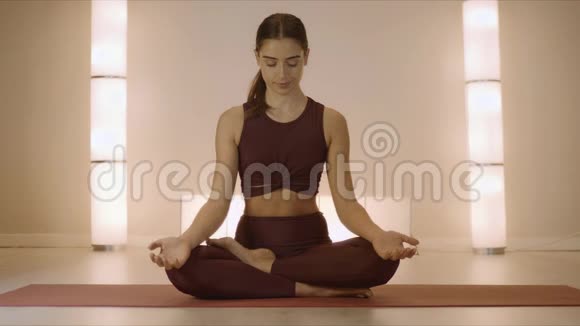 女人坐在莲花的姿势在室内的垫子上教练在工作室练习瑜伽视频的预览图