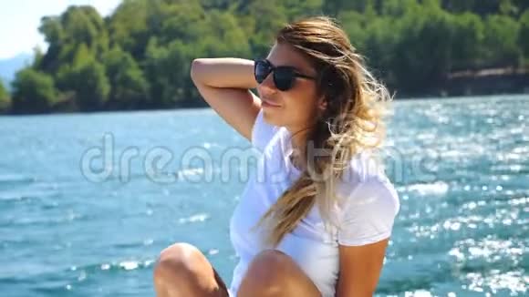 快乐的金发女郎坐在游艇上在风景优美的景色下漂浮在海湾戴太阳镜的年轻女孩视频的预览图
