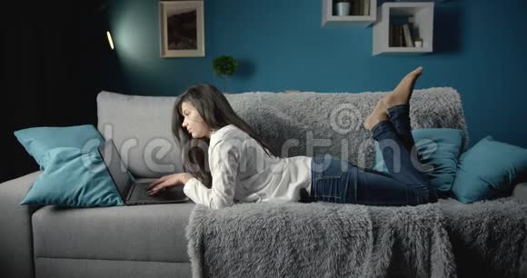 穿休闲服装的女人躺在沙发上用笔记本电脑视频的预览图