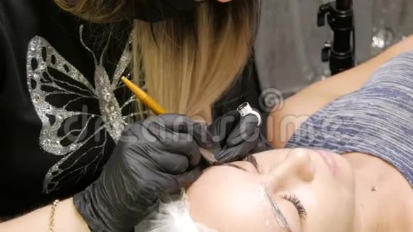 戴黑色手套的美容师为年轻漂亮的女孩做永久性的化妆矫正眉毛的形状视频的预览图