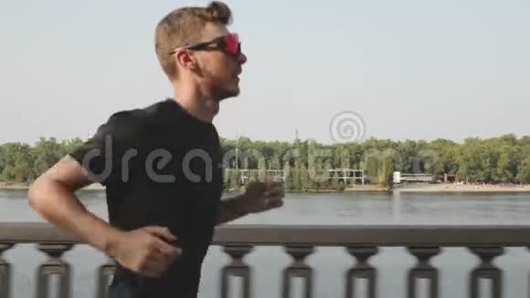 强壮的年轻人穿着黑色t恤和运动太阳镜沿河慢跑精力充沛的活跃男孩跑步性感男人训练视频的预览图