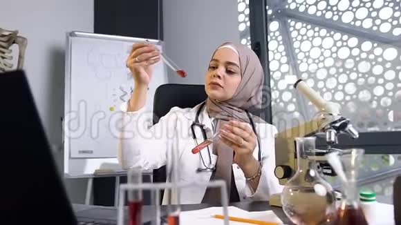 有魅力的35岁穆斯林女医生戴着头巾在诊所的两个试管中比较化学液体视频的预览图