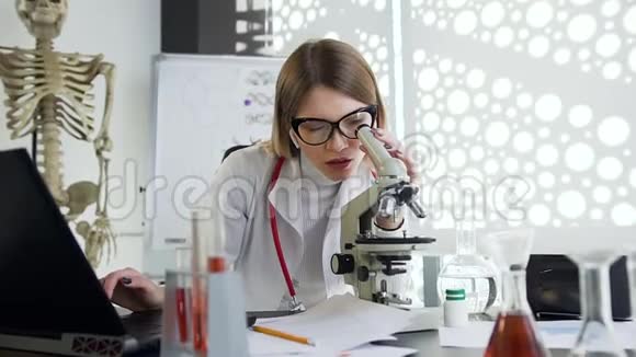 带着显微镜和电脑在诊所的橱柜里工作的漂亮愉快的年轻医生视频的预览图