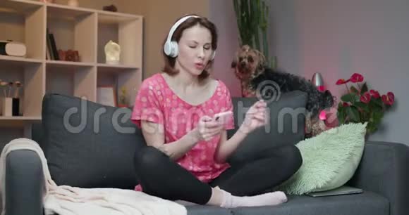 白人女孩坐在家里的沙发上微笑着在现代智能手机上用耳机听音乐视频的预览图