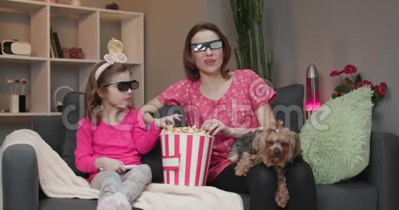 女人带着一个小女孩和狗戴着3D眼镜看电视和吃爆米花家庭时间放松年轻女孩视频的预览图