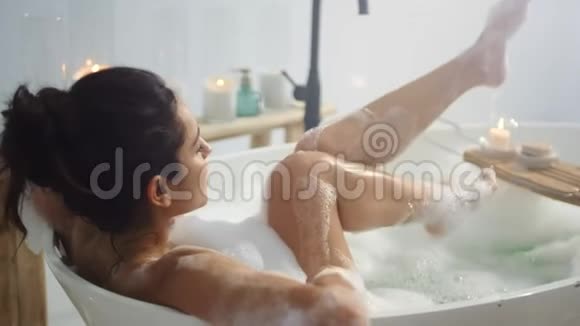 放松的女人在浴缸里挥舞着腿从侧面看深褐色头发的女孩洗腿视频的预览图