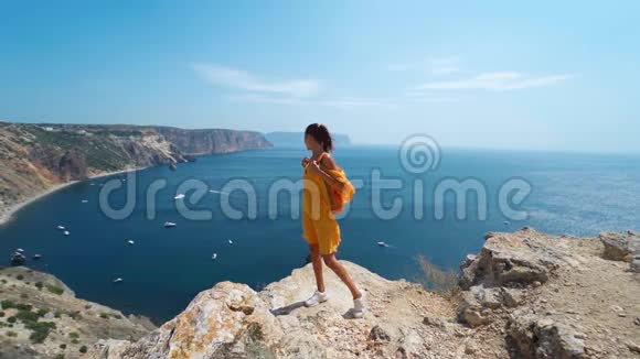 黝黑的旅人女人站在悬崖边穿着明亮的黄色连衣裙在风中飘扬视频的预览图