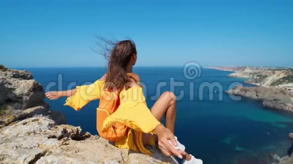 特写一幅晒黑了的旅行妇女的肖像她举起双手坐在岩石上的海滩上穿着鲜艳的黄色连衣裙和头发视频的预览图