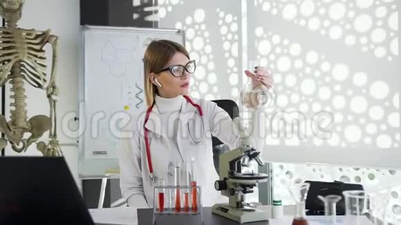 美丽的金发女医生坐在桌子旁用试管和装有化学物质的烧瓶工作视频的预览图