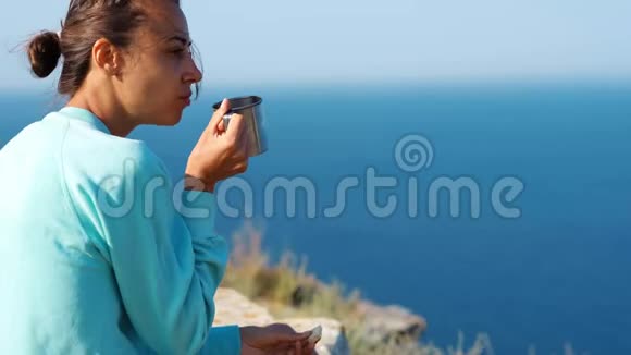 户外肖像无忧无虑的微笑女人坐在悬崖边美丽的海景拿着杯子咖啡或茶视频的预览图