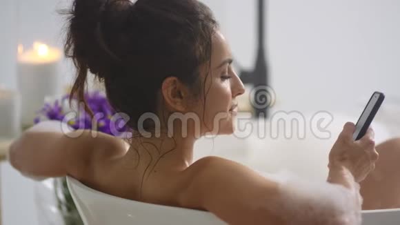 可爱的女孩在浴缸里的智能手机上打字性格开朗的女人打电话放松视频的预览图