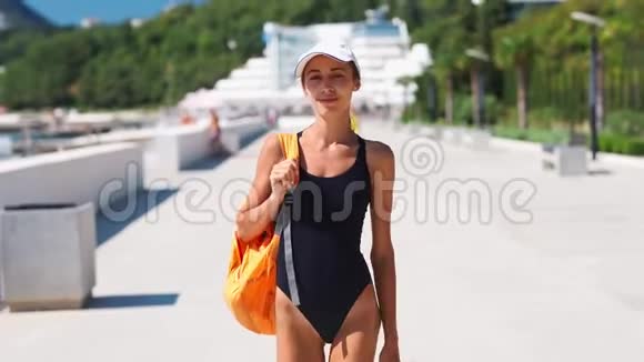 穿着黑色泳装白色帽子和橙色背包的漂亮时尚性感女士漫步在海边的海湾视频的预览图