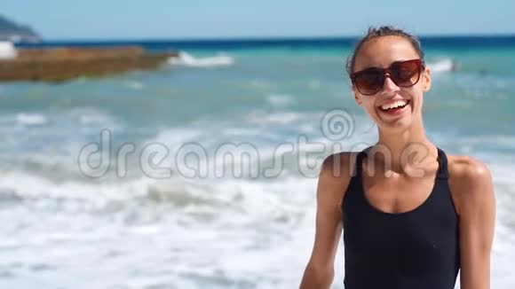 快乐地笑着性感苗条的女孩穿着黑色泳装戴着太阳镜站在美丽的阳光明媚的海滩上视频的预览图