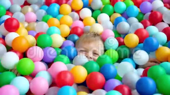 金发小男孩躺在多种颜色的塑料球在大干划水池在游戏中心对着镜头微笑视频的预览图