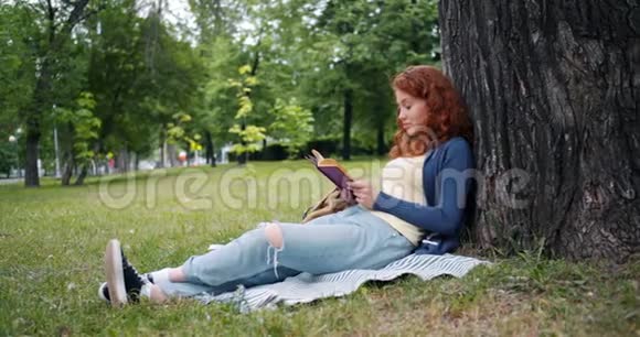 美丽的年轻女子坐在公园树下的毯子上看书微笑着视频的预览图