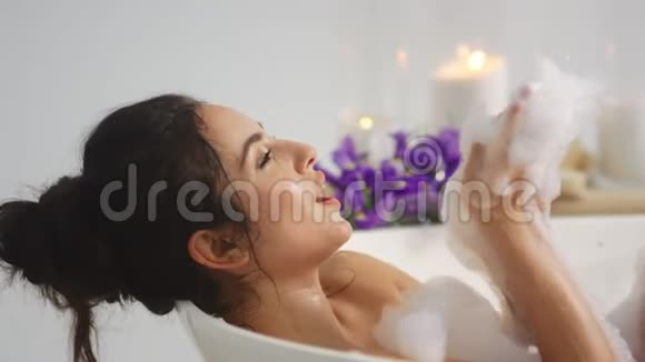 侧面看放松的女人玩泡沫在浴缸里微笑的漂亮女人视频的预览图