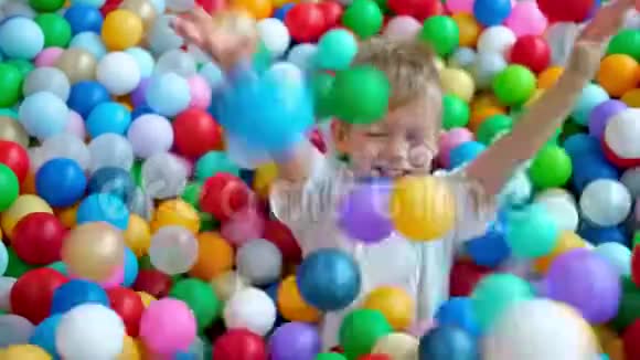 金发的小男孩把球分散在不同的侧面躺在多种颜色的塑料球上在大干划池里视频的预览图