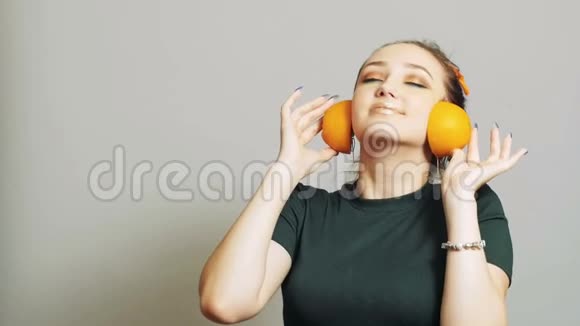 漂亮的女孩把橘子的一半水果放进耳朵里就像耳机一样青少年在演播室背景上玩得很开心概念音乐视频的预览图