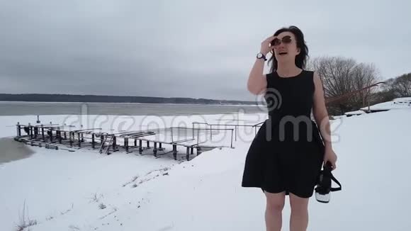 穿着黑色无雪裙的布鲁内特站在雪覆盖的河岸上带着她的太阳镜觉得冷然后视频的预览图