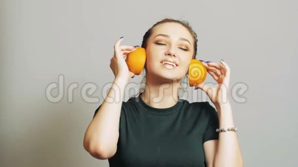 漂亮的女孩把橘子的一半水果放进耳朵里就像耳机一样青少年在演播室背景上玩得很开心概念音乐视频的预览图