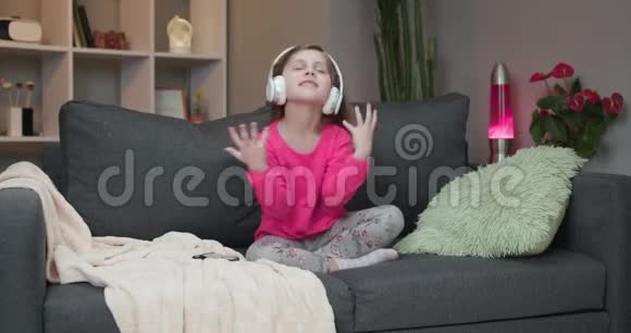 小女孩在沙发上跳舞在家听耳机里的音乐戴着耳机跳舞唱歌唱歌的女孩视频的预览图
