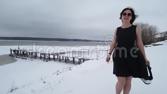 穿着黑色连衣裙和太阳镜的女人在覆盖着雪的河岸上跳跃和跳舞使自己温暖正面观点视频的预览图